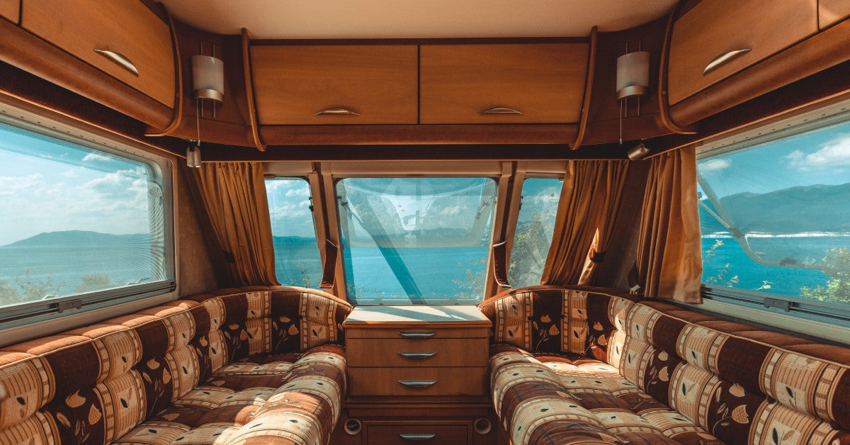 A view from inside a caravan features an extended couch along two walls and a wooden drawer at the end.