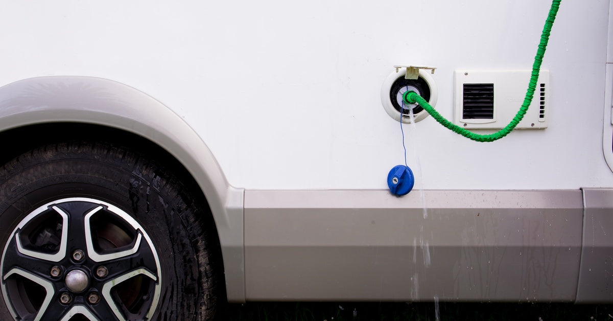 A close-up of an RV's side with a green hose connected to it. Water drips onto wet grass near the tire.