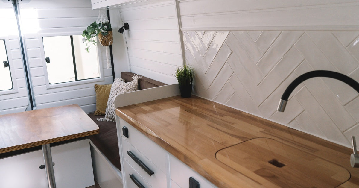 The interior of a motorhome with an emphasis on the kitchen. There are wooden countertops with a board over the sink.