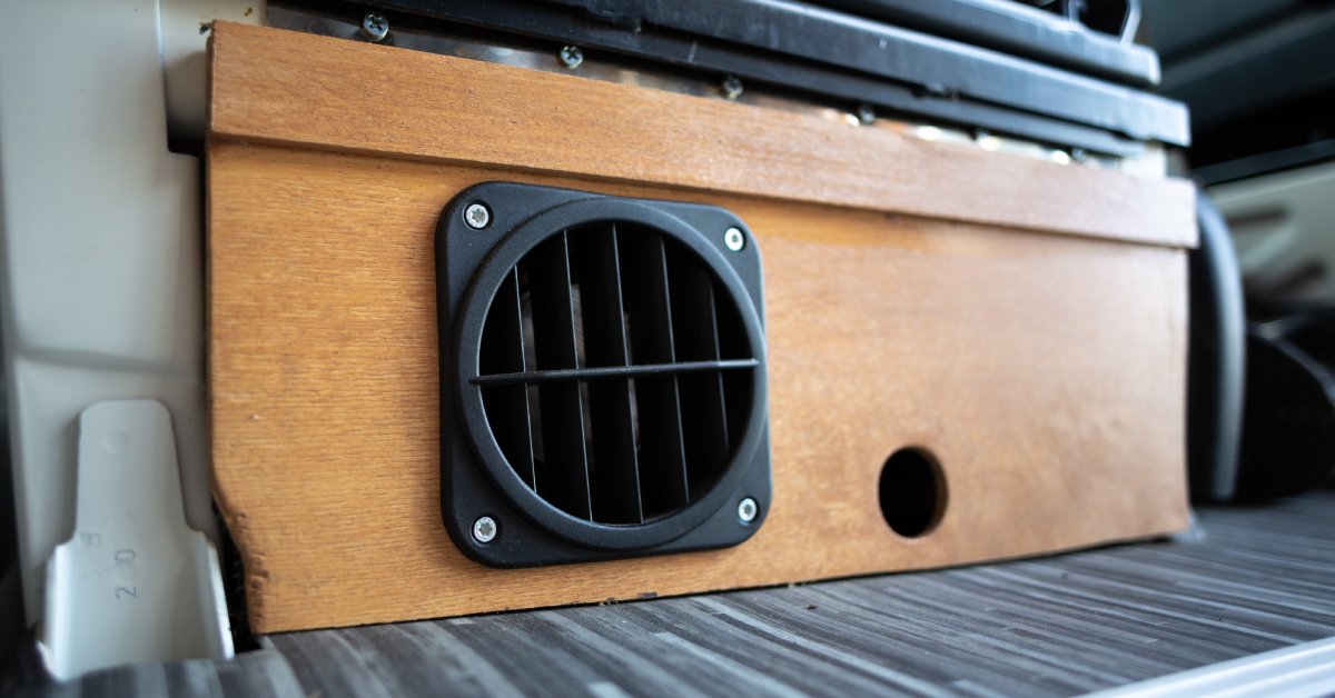 A round black heating vent on a wooden panel in an RV. The heat comes from under the driver's seat.