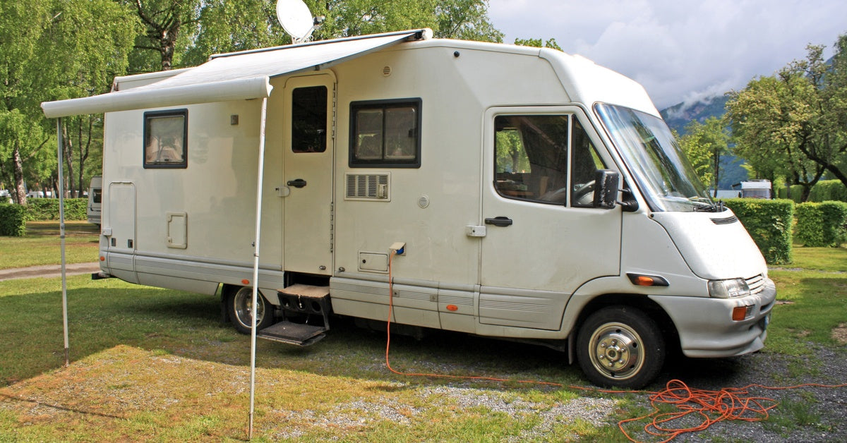 A white camper van parked in a grassy field with wildflowers. A white cloth awning stretches out above the side door.