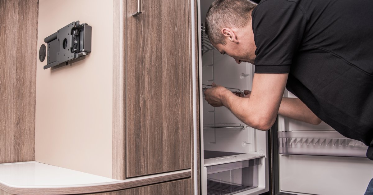 A man in a black T-shirt is standing in front of an open RV refrigerator. He's reaching inside and grabbing a shelf.