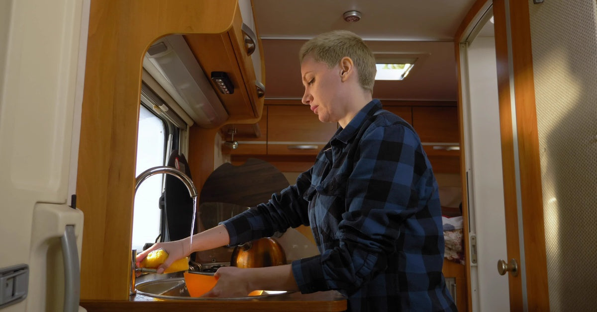 A person with short hair and a blue plaid shirt is standing at the kitchen sink of an RV. A small stream of water flows from it.