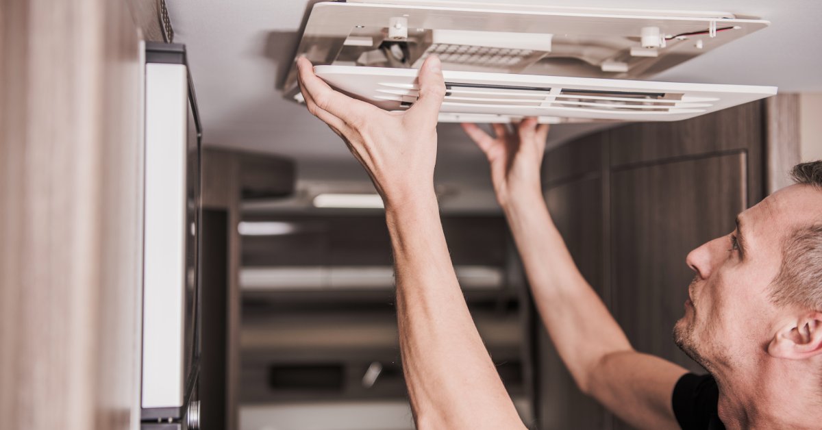 A man in a black T-shirt is standing in an RV. He's carefully lifting the panel off of a ceiling-mounted A/C unit.