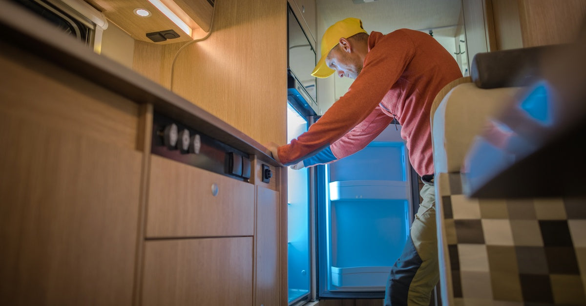 A man wearing an orange sweatshirt and a yellow baseball cap leans on wooden furniture, looking inside an open fridge.