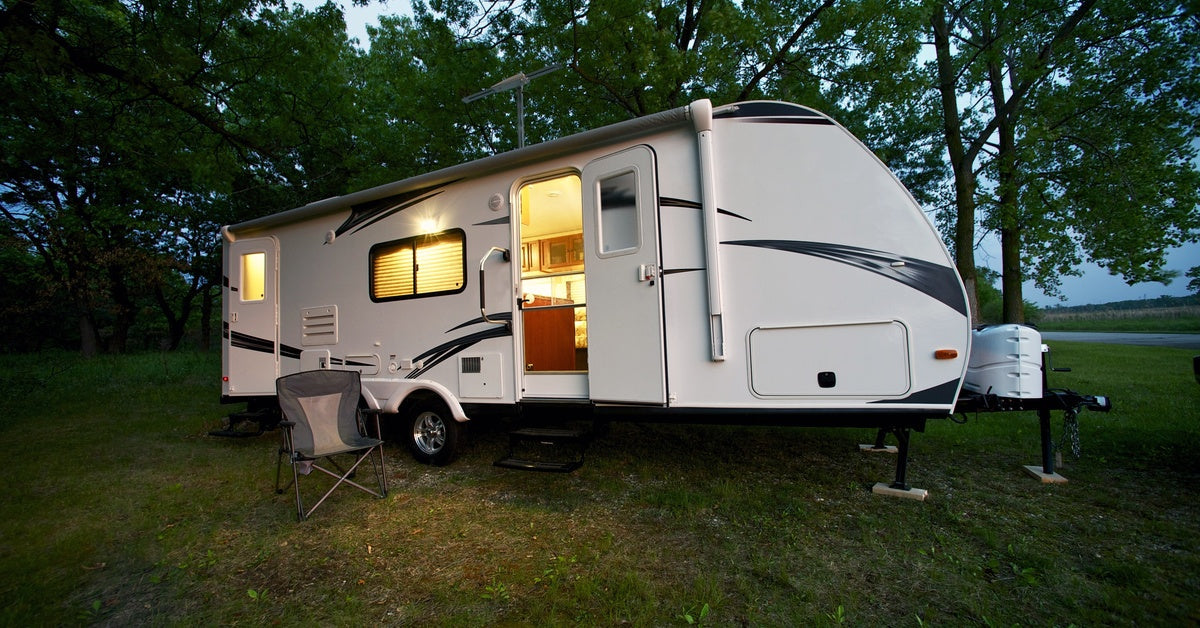 A camper is parked on a grassy yard with a chair outside. The front door is open and all the lights are on inside.